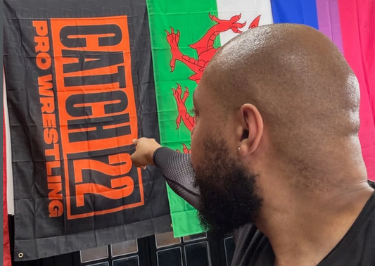 Person holding a pro wrestling banner with 'Catch 22' branding in front of a colorful flag.